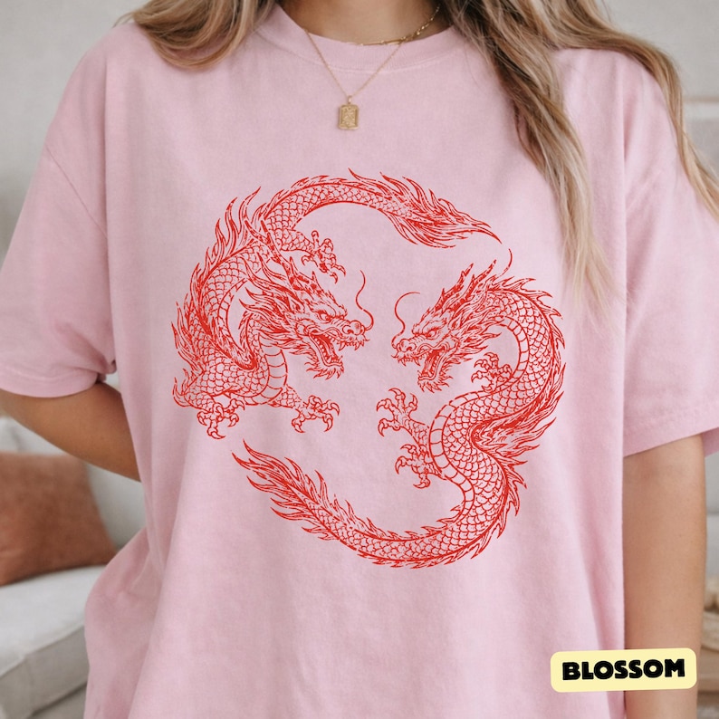 May include: A light pink short-sleeved t-shirt featuring a large, detailed red graphic of two dragons intertwined in a circular pattern. The design is centred on the chest. A small rectangular label with the text BLOSSOM is in the bottom corner.