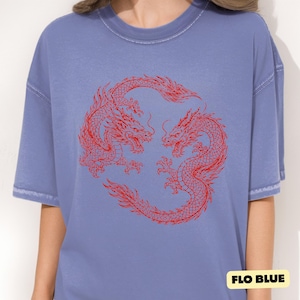 May include: A periwinkle blue short-sleeved t-shirt featuring a circular graphic of two red dragons facing each other in a mirrored pose. The fabric has a slightly faded, washed-out appearance. A small yellow label in the corner reads FLO BLUE.