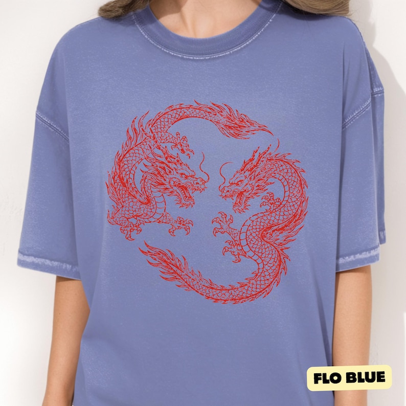 May include: A periwinkle blue short-sleeved t-shirt featuring a circular graphic of two red dragons facing each other in a mirrored pose. The fabric has a slightly faded, washed-out appearance. A small yellow label in the corner reads FLO BLUE.