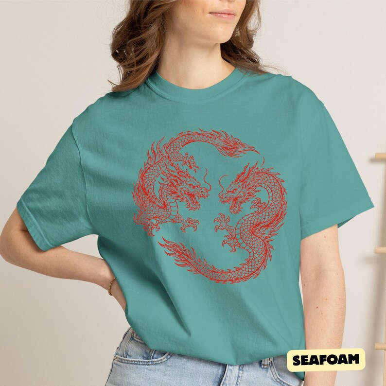 May include: A seafoam green short-sleeved t-shirt featuring a circular graphic of two red dragons facing each other in a stylised design. The shirt has a crew neckline and is paired with blue denim jeans. The text "SEAFOAM" is printed in the corner.