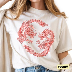 May include: An ivory short-sleeved t-shirt featuring a circular graphic of two red dragons facing each other in a mirrored pose. The dragons have detailed scales, flowing manes, and sharp claws. The word IVORY is printed in a small black box at the bottom.
