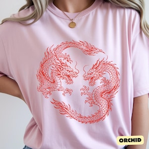 May include: A light pink short-sleeved t-shirt featuring a circular graphic of two red dragons facing each other. The dragons are depicted with detailed scales, flowing manes, and long tails, forming a ring shape in the centre of the garment.