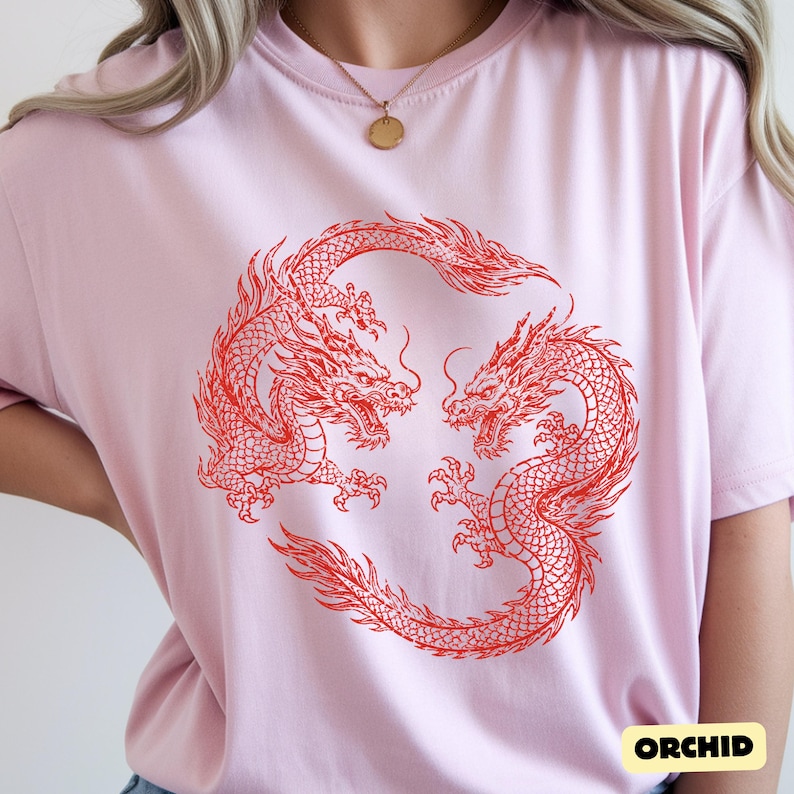 May include: A light pink short-sleeved t-shirt featuring a circular graphic of two red dragons facing each other. The dragons are depicted with detailed scales, flowing manes, and long tails, forming a ring shape in the centre of the garment.