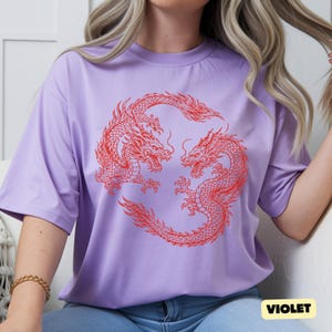 May include: A light purple short-sleeved t-shirt featuring a circular graphic of two red dragons facing each other. The dragons are detailed with scales and flowing whiskers, arranged in a symmetrical pattern on the centre of the chest area.