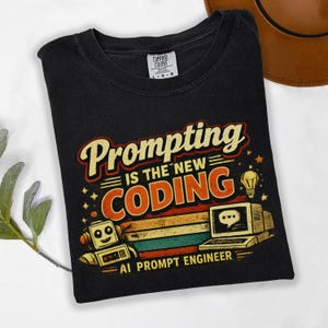 Op de afbeelding: Zwart t-shirt met de tekst "Prompting is the new Coding" in retro-ontwerp. De afbeelding bevat een robot, computer en gloeilamp, met de woorden "AI Prompt Engineer" eronder. Comfort Colors merk.