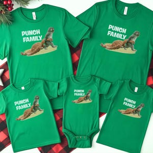 Könnte beinhalten: Grüne T-Shirts und ein Baby-Body mit dem Text "PUNCH FAMILY" und einer Grafik von zwei Tieren. Die Shirts sind auf einer rot-schwarz karierten Oberfläche mit einem Zweig Grünzeug in der oberen linken Ecke angeordnet.