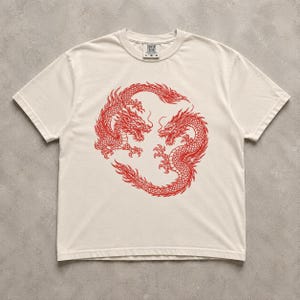 May include: A cream-coloured short-sleeve t-shirt featuring a large, detailed red graphic of two dragons intertwined in a circular pattern on the chest. The shirt has a crew neckline and a visible brand label at the inner collar reading Comfort Colors.
