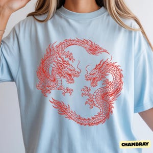 May include: A light blue short-sleeved t-shirt featuring a large, detailed red graphic of two dragons arranged in a circular, interlocking pattern. The dragons are depicted with intricate scales, flowing manes, and sharp claws, facing each other.