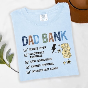 May include: Light blue t-shirt with the words "DAD BANK" in colorful letters. The shirt features a checklist with phrases like "ALWAYS OPEN" and "INTEREST-FREE LOANS", along with a lightning bolt and coins.