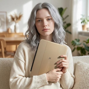 Peut inclure: Une housse de tablette beige avec les initiales "JK" en noir est tenue par une personne portant un pull crème. L'étui a une ligne verticale au centre. La personne a les cheveux gris et est assise sur un canapé beige.