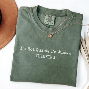 May include: An olive green t-shirt with the text "I'm Not Quiet, I'm Just... THINKING" in white. A gold-colored necklace with a circular pendant rests on the shirt. The shirt has a Comfort Colors tag.