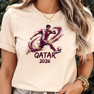 A woman wearing a white t-shirt with a graphic of a soccer player and the text "Qatar 2026" printed on it.