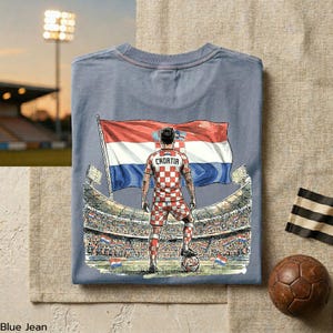 A t-shirt with a graphic design depicting a soccer player standing in front of a stadium, holding a soccer ball and wearing a red and white checkered jersey with the name 'Croatia' on the back.