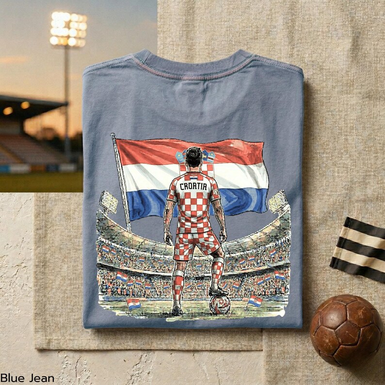 A t-shirt with a graphic design depicting a soccer player standing in front of a stadium, holding a soccer ball and wearing a red and white checkered jersey with the name 'Croatia' on the back.