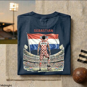 A t-shirt design featuring a soccer player standing in front of a stadium with a large flag, with the text "SEBASTIAN" and "CROATIA" printed on it.