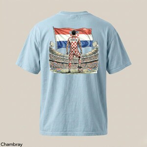 A light blue t-shirt with a graphic design depicting a soccer player standing in front of a stadium, holding a flag with the colors of the Croatian national flag.