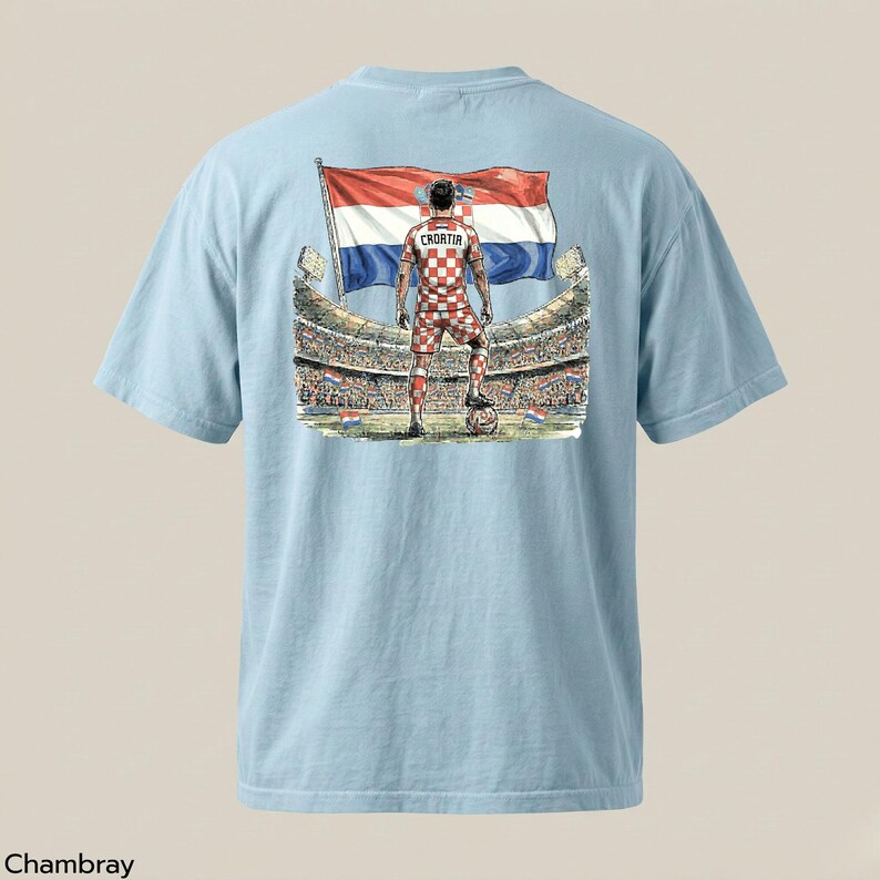 A light blue t-shirt with a graphic design depicting a soccer player standing in front of a stadium, holding a flag with the colors of the Croatian national flag.