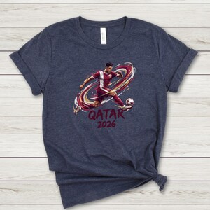 A dark blue t-shirt with a graphic design depicting a soccer player kicking a ball, along with the text "QATAR 2026" printed on it.