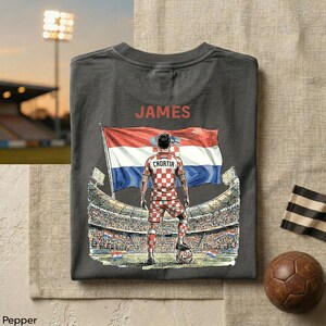 A gray t-shirt with a graphic design depicting a soccer player in a red and white checkered uniform, standing in front of a stadium with a flag, and the name 'JAMES' printed on the back.