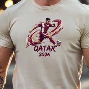 A person wearing a white t-shirt with a graphic design depicting a soccer player in a red uniform kicking a soccer ball.