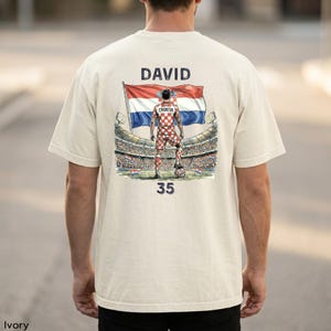 A person wearing a white t-shirt with a graphic design depicting a soccer player standing in front of a stadium, holding a flag.