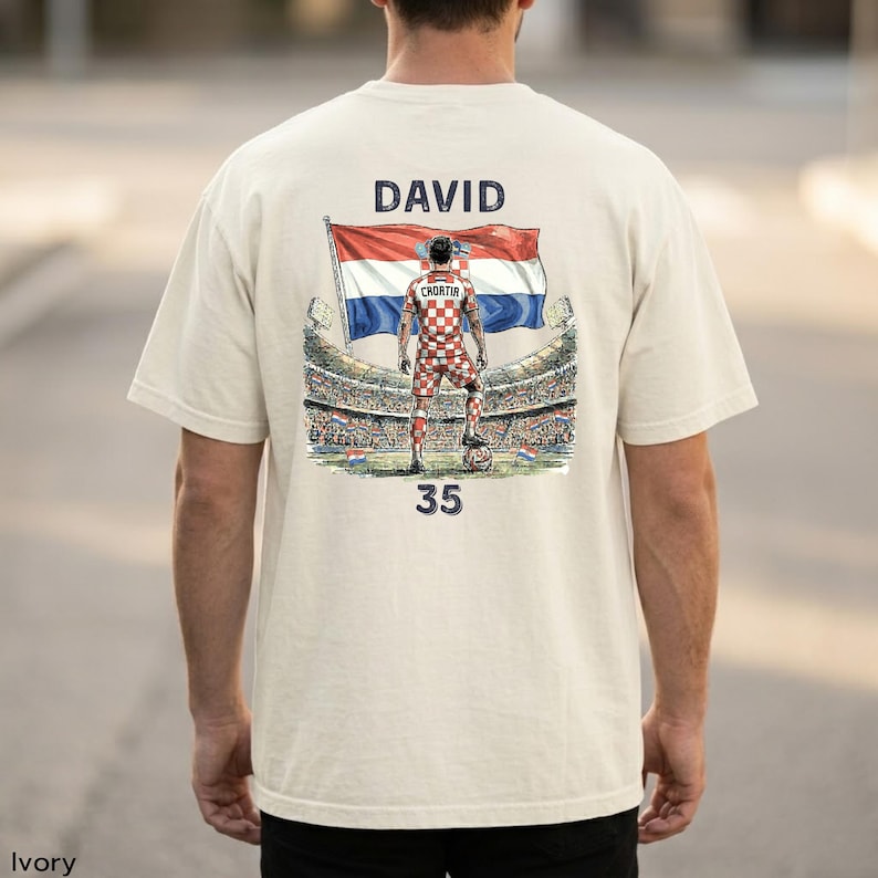 A person wearing a white t-shirt with a graphic design depicting a soccer player standing in front of a stadium, holding a flag.