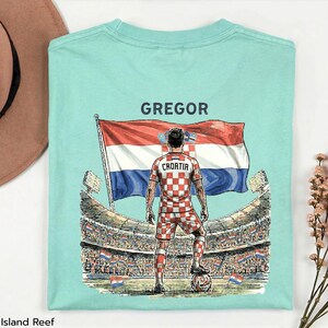 A t-shirt design featuring a soccer player in a red and white checkered uniform standing in front of a large crowd at a soccer stadium, with the word "GREGOR" written above the player.
