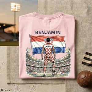 A pink t-shirt with a graphic design depicting a soccer player standing in front of a stadium, holding a flag, and the name "BENJAMIN" printed on it.