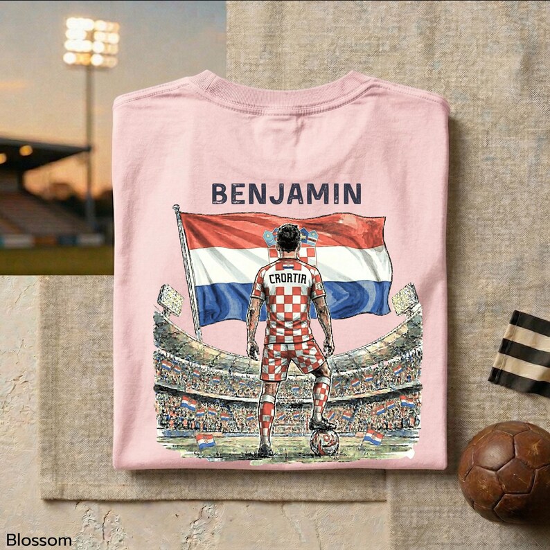 A pink t-shirt with a graphic design depicting a soccer player standing in front of a stadium, holding a flag, and the name "BENJAMIN" printed on it.