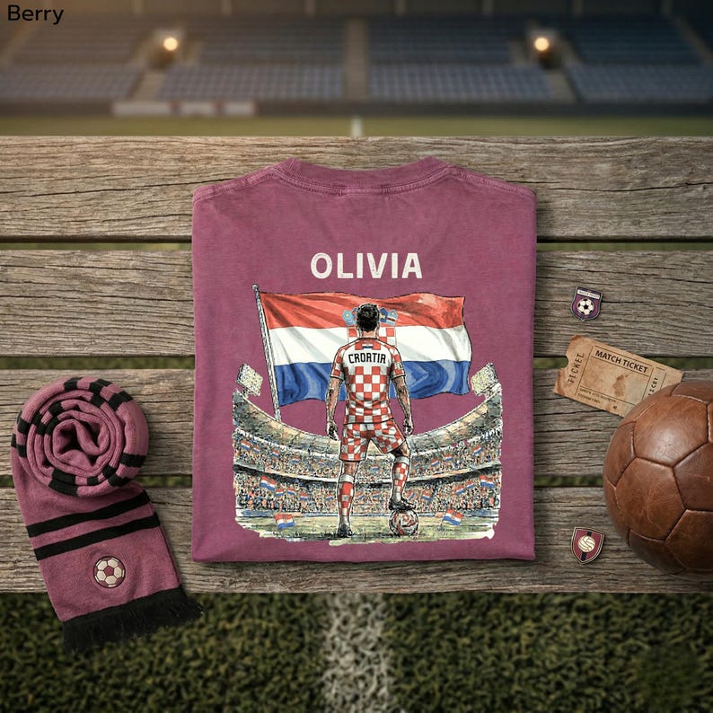 a t-shirt with a graphic of a soccer player, a soccer ball, a scarf, and some other sports-related items arranged on a wooden surface.