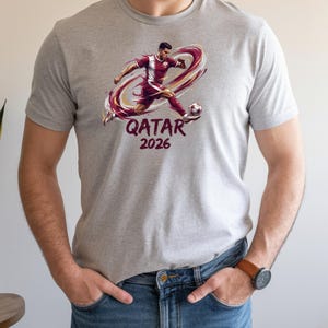 A man wearing a grey t-shirt with a graphic of a soccer player and the text "Qatar 2026" printed on it.