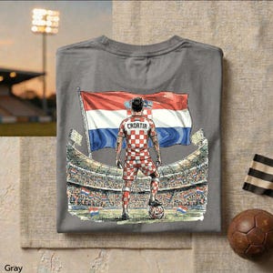 A gray t-shirt with a graphic design depicting a soccer player standing in front of a large crowd, holding a soccer ball and wearing a red and white checkered jersey with the word "Croatia" on the back.