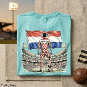 A t-shirt with a graphic design depicting a soccer player standing in front of a stadium, holding a flag, and wearing a checkered jersey.