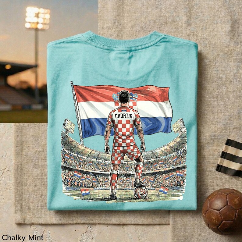 A t-shirt with a graphic design depicting a soccer player standing in front of a stadium, holding a flag, and wearing a checkered jersey.
