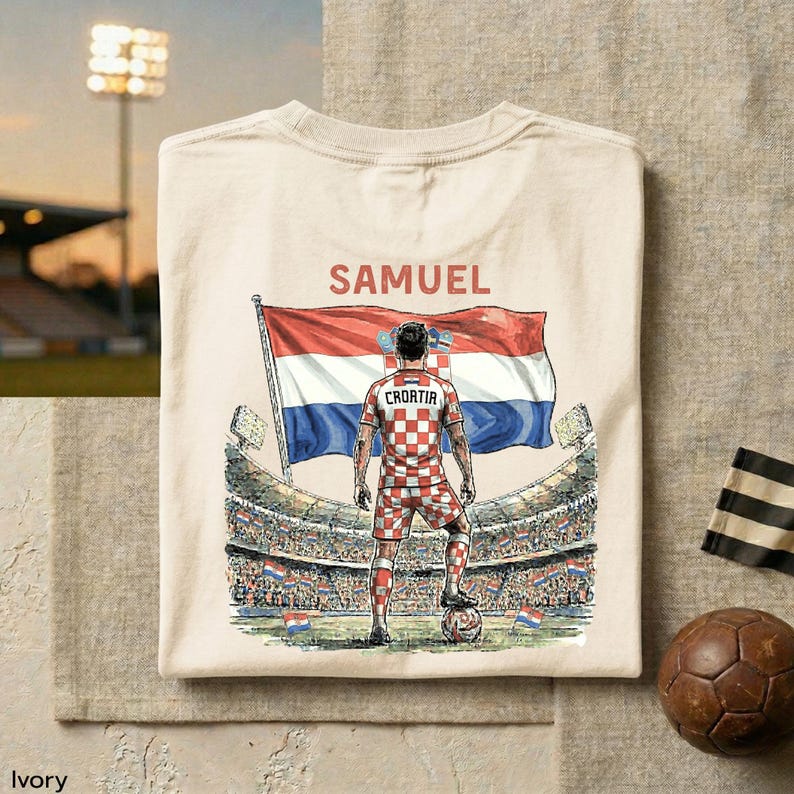 A white t-shirt with a graphic design depicting a soccer player standing in front of a stadium, holding a soccer ball and wearing a red and white checkered jersey.