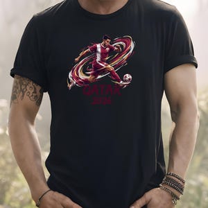 A person wearing a black t-shirt with a graphic of a soccer player kicking a ball, along with the text "QATAR 2026" printed on it.