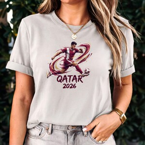 A woman wearing a white t-shirt with a graphic of a soccer player and the text "Qatar 2026" printed on it.