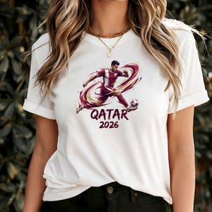 A woman wearing a white t-shirt with a graphic of a soccer player and the text "QATAR 2026" printed on it.
