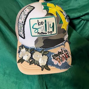 以下が含まれることがあります： ベージュと迷彩柄のトラッカーハット。花柄と刺繍のパッチが付いています。「be salty Matthew 5:13」と「Powered by Prayer」の文字が特徴です。黄色の十字架と白い花が帽子を飾っています。