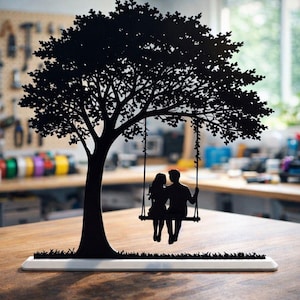 May include: Black silhouette of a tree with a swing, featuring a couple. The tree is on a white base with a grass design. The background is a workshop with tools and equipment. A decorative item.