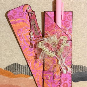 May include: A pink and orange handmade bookmark with a matching pen holder. The bookmark has a tag that says "Timeless". A decorative butterfly and embellishments adorn the pen holder. The background has a landscape design.