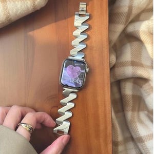 Puede incluir: Reloj inteligente plateado con una correa metálica ondulada única. La esfera muestra un corazón morado y la hora, 12:43. El reloj está sobre una superficie de madera. La correa mide unos 18 cm de largo.