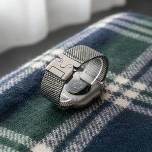 Può includere: Uno smartwatch argentato con cinturino a rete e chiusura di sicurezza. L'orologio è esposto su una superficie a fantasia a quadri verdi e blu. Il cinturino è realizzato in materiale tessuto.