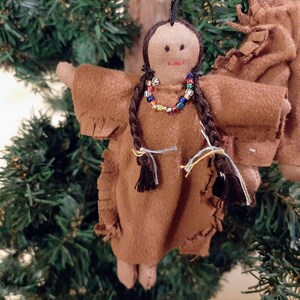 May include: A brown felt doll with long black braids, wearing a brown dress with fringe and a colorful beaded necklace. The doll is hanging from a Christmas tree.