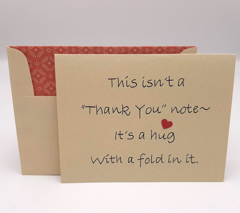 以下が含まれることがあります： 「This isn't a "Thank You" note~ It's a hug with a fold in it.」と書かれた茶色のカードと小さな赤いハート。