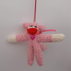 May include: A handmade sock monkey ornament in pink and white knit. The monkey has a red mouth, black eyes, and a pink heart-shaped button on its chest. A pink ribbon is tied around its neck, and a red string is attached for hanging.
