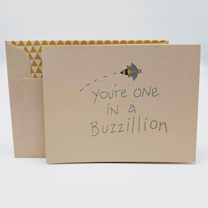 May include: A light pink card with a yellow and white patterned envelope. The card has a black and gray illustration of a bee flying with the text "you're one in a Buzzillion".