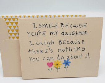 May include: A card with a handwritten message that reads "I smile because you're my daughter. I laugh because there's nothing you can do about it." The card has a yellow, white, and blue floral design.