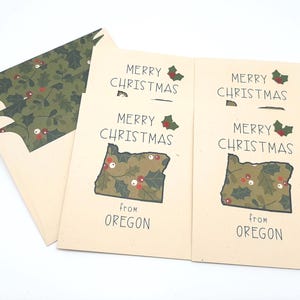 May include: Christmas cards with a cream-colored background. The cards feature the text "Merry Christmas" and a map of Oregon filled with a holly and berry pattern. An envelope with a matching holly pattern is also visible.