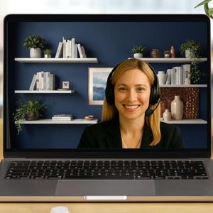 May include: A laptop displaying a smiling woman wearing a headset, likely in a video call. The screen shows a blue wall with shelves, books, and plants. The laptop sits on a wooden desk with office supplies and a potted plant.